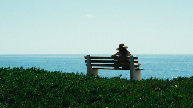 a woman spending time alone sitting on a bench on the grass staring at the beautiful blue sea