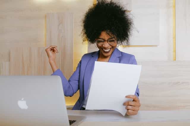 woman in office with blazer and funky hair wearing glasses holding paper in her hands and smiling at them with her mac on the desk