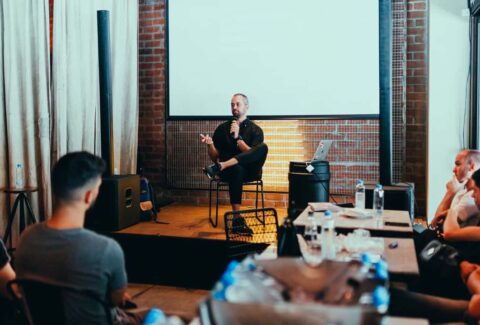 a guy sitting down in front of a crowd holding mic speaking with a white board behind him