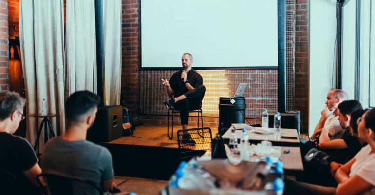 a guy sitting down in front of a crowd holding mic speaking with a white board behind him