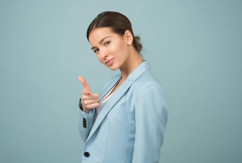 a woman in a light blue blazer feeling confident smiling at the camera and pointing her index finger at the camera with blue background behind her