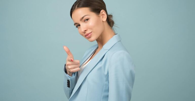 a woman in a light blue blazer feeling confident smiling at the camera and pointing her index finger at the camera with blue background behind her