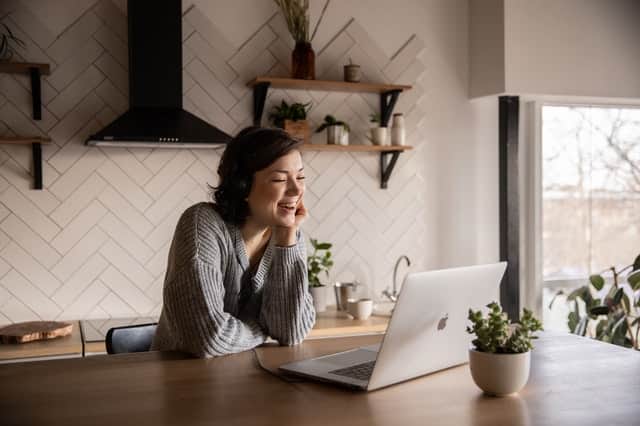 Woman smiling at her laptop on her desk in her kitchen with a small plant next to her