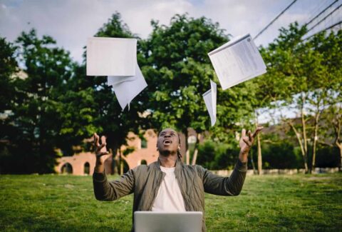 a guy in the park throwing his paper documents in the air with his laptop in front of him smiling in excitement