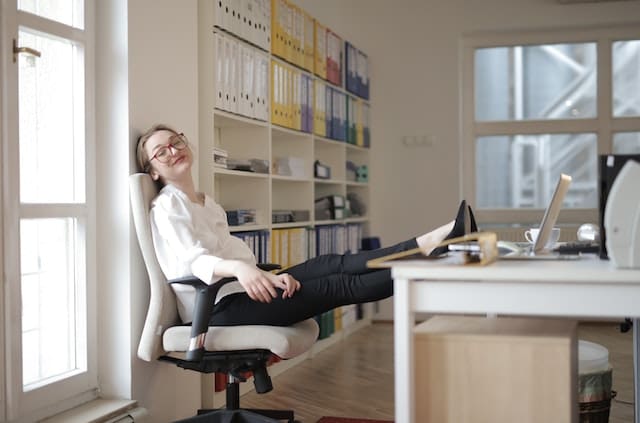 A woman feeling happy and fulfilled at work with her rewarding job. She is smiling on her chair, legs up on her desk next to her laptop.