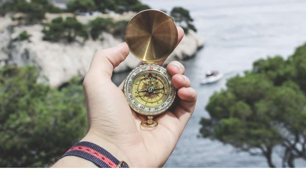 A guy holding a compass in nature overlooking the sea and trees 