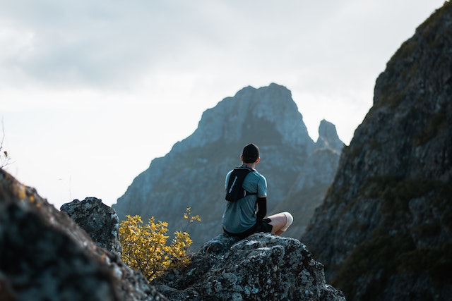 a guy sitting on top of a mountain rock staring at other mountains 