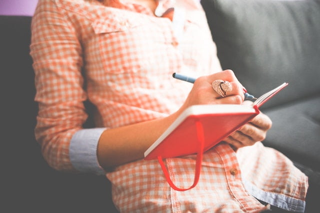 a woman wearing a read and white checked shirt sitting on a sofa with a journal and pen in her hands practicing journaling to break codependency