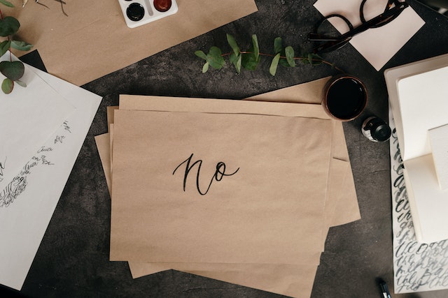 a brown paper with the word 'no' hand-written on it on a desk with papers, green plants, and glasses 