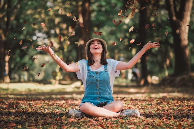 a girl sitting on the floor in a forest on autumn leaves with her hands wide open as she throws leaves in the air, she looks up at them and smiles feeling alive