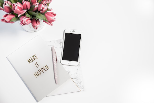 a flower arrangement next to a journal written on it "MAKE IT HAPPEN" with a pen on it and a mobile. 