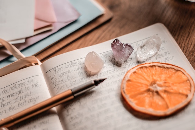 A journal with crystals on it, a slice of orange and a pen. Journaling is a technique used in life purpose coaching.