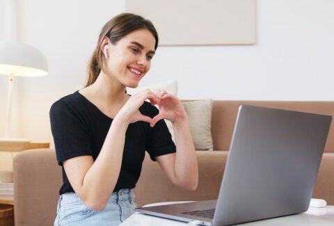 a girl on an online call on her laptop doing life purpose coaching and smiling making a love heart symbol with her arms