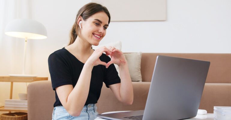 a girl on an online call on her laptop doing life purpose coaching and smiling making a love heart symbol with her arms