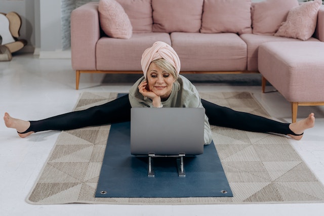 a 50 year old woman rediscovering her passions on the floor in her living room on her yoga mat looking at the laptop smiling legs in split form with her bath towel on her head 