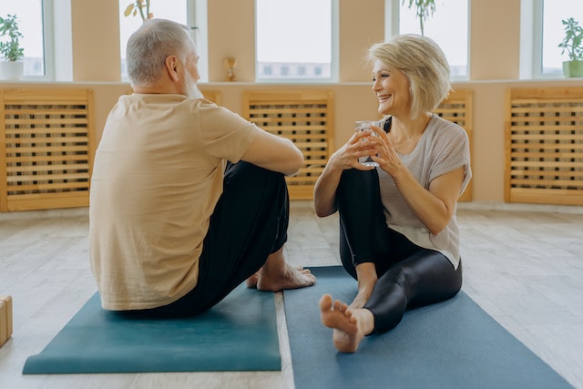 older adults male and female sitting on the floor on their yoga mat having a healthy conversation and smiling