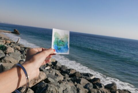 what is spiritual counseling - a girl holding a paper in her hand written 'I am peace' on the rocks with the beautiful blue sea behind