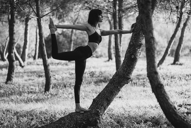 a woman in a forest doing yoga reaching out to her leg while touching a tree