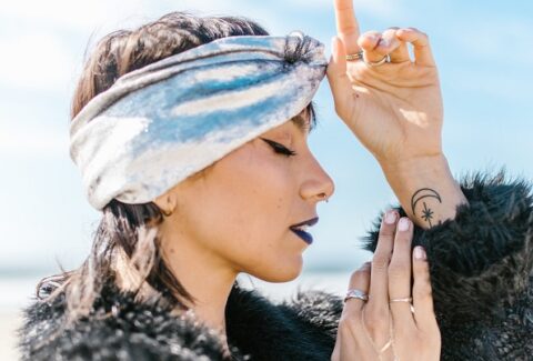 A woman at the beach wearing a chic turban with eyes closed, hands on third eye, wearing winged eye liner, moon and stars tattoo embracing her spiritual awakening