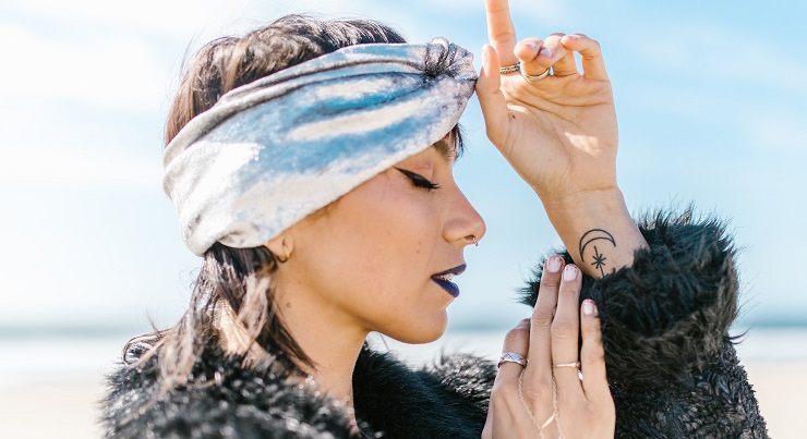 A woman at the beach wearing a chic turban with eyes closed, hands on third eye, wearing winged eye liner, moon and stars tattoo embracing her spiritual awakening