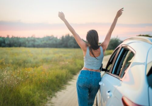 a girl solo traveling in a field with green grass next to her car putting her hands up with the sky with joy, signs you're healing from narcissistic abuse