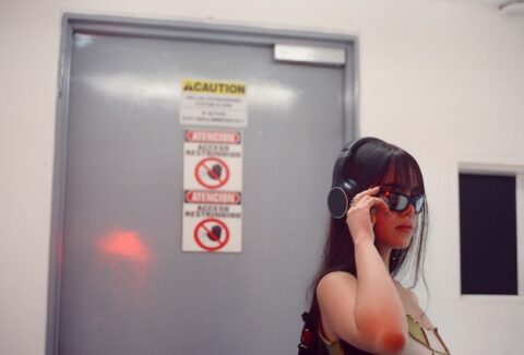 a woman wearing headphones and sunglasses in a white room with a door behind her with warning signs of caution written on the door behind her