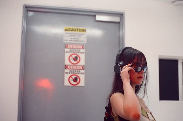 a woman wearing headphones and sunglasses in a white room with a door behind her with warning signs of caution written on the door behind her