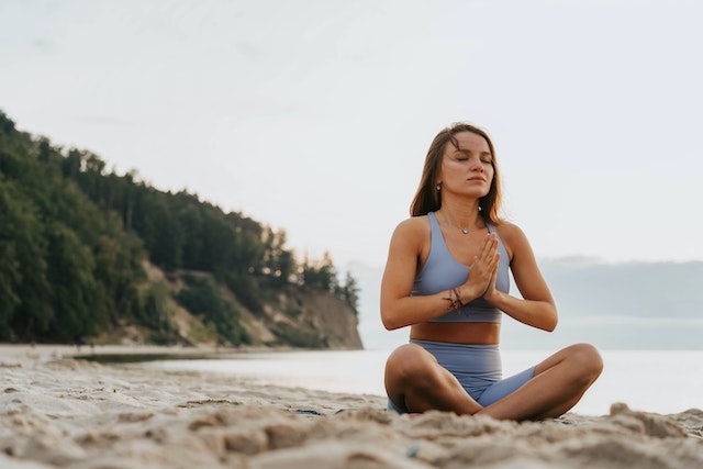 a woman on the sand next to the sea with green hills behind her in lotus prayer position eyes closed