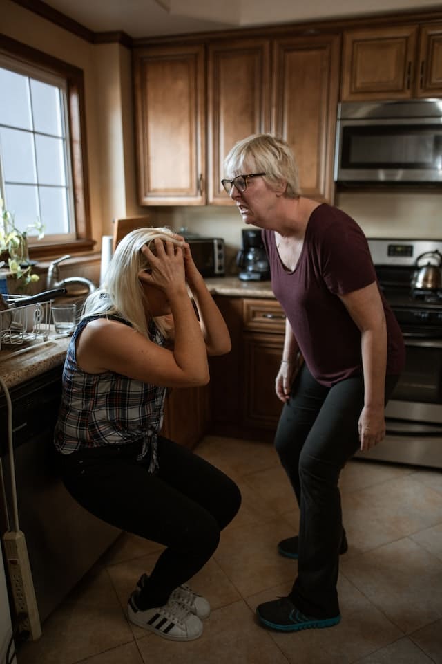 narcissistic parent verbally abusing her daughter in the kitchen as her daughter covers her face with her hands in despair