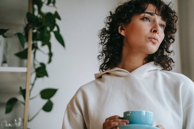 a woman staring blankly outside the window holding a mug in her hand