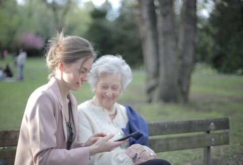 an old woman on a bench sitting in the park next to her adult daughter looking at her phone