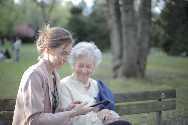 an old woman on a bench sitting in the park next to her adult daughter looking at her phone